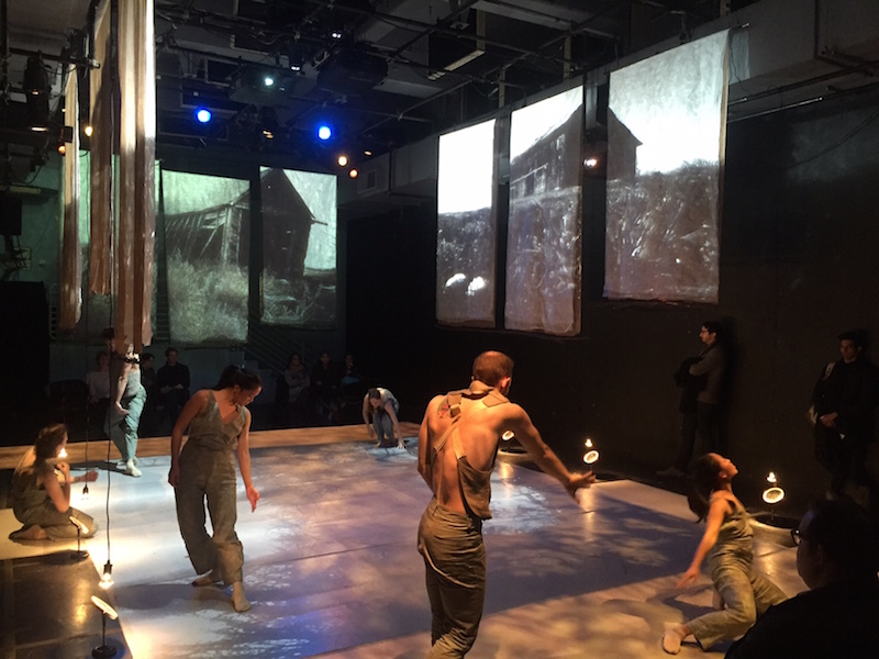 Dancers in overalls and jumpers dancing on the perimeter of the stage with projections of old homes and barns hanging from the ceiling